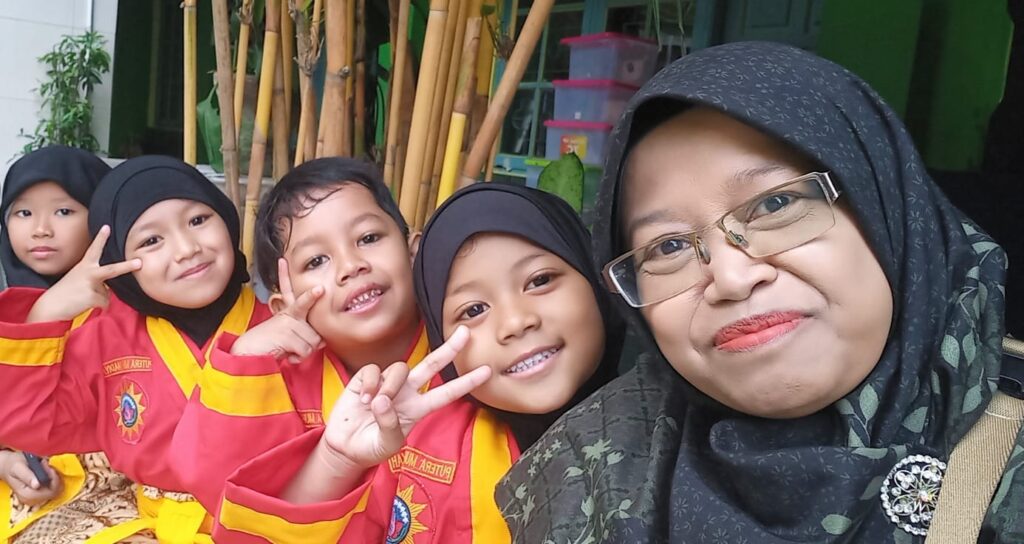 Teacher in hijab and glasses taking a selfie with four smiling children in red and yellow uniforms outdoors
